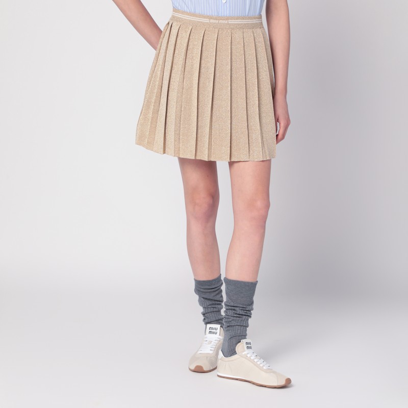 플리츠 디자인, 신축성 있는 리브 허리띠, 대조적인 앞면 로고 인레이가 특징인 Miu Miu의 골드 루렉스 미니 스커트입니다. 이미지