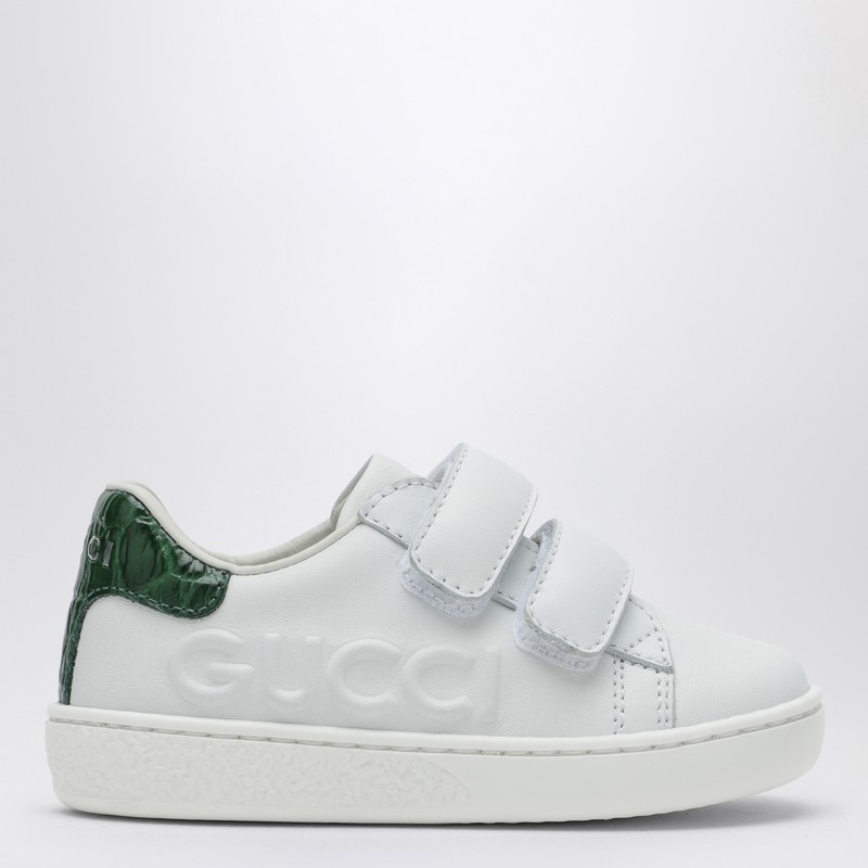 구찌의 흰색 가죽 로우탑 스니커즈는 둥근 앞부분, 벨크로 패스너, 뒷면의 녹색 로고 디테일, 옆면의 양각 문자, 고무 밑창이 특징입니다. 이미지