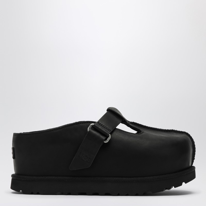 검은색 가죽으로 만든 Ugg 클로그는 둥근 앞부분, 내부 플랫폼 밑창, 벨크로 잠금장치와 로고가 있는 스트랩이 있는 윗부분, 뒷면 로고 패치와 고무 밑창이 특징입니다. 이미지