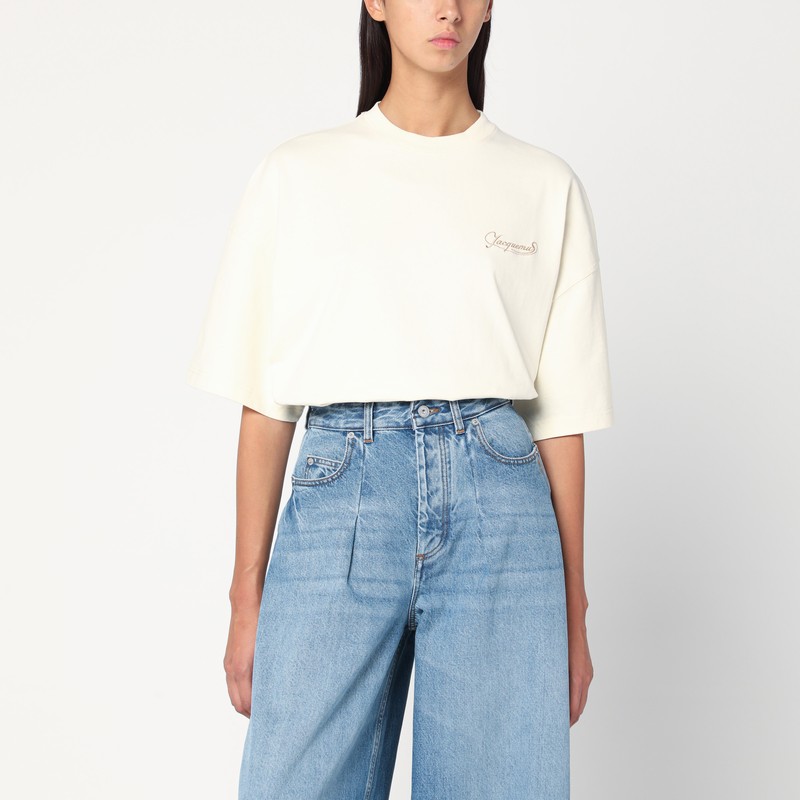 자크뮈스의 밝은 베이지색 코튼 티셔츠는 크롭과 오버사이즈 핏, 리브 크루넥, 가슴에 대조적인 자수 로고 레터링, 3/4 소매가 특징입니다. 이미지