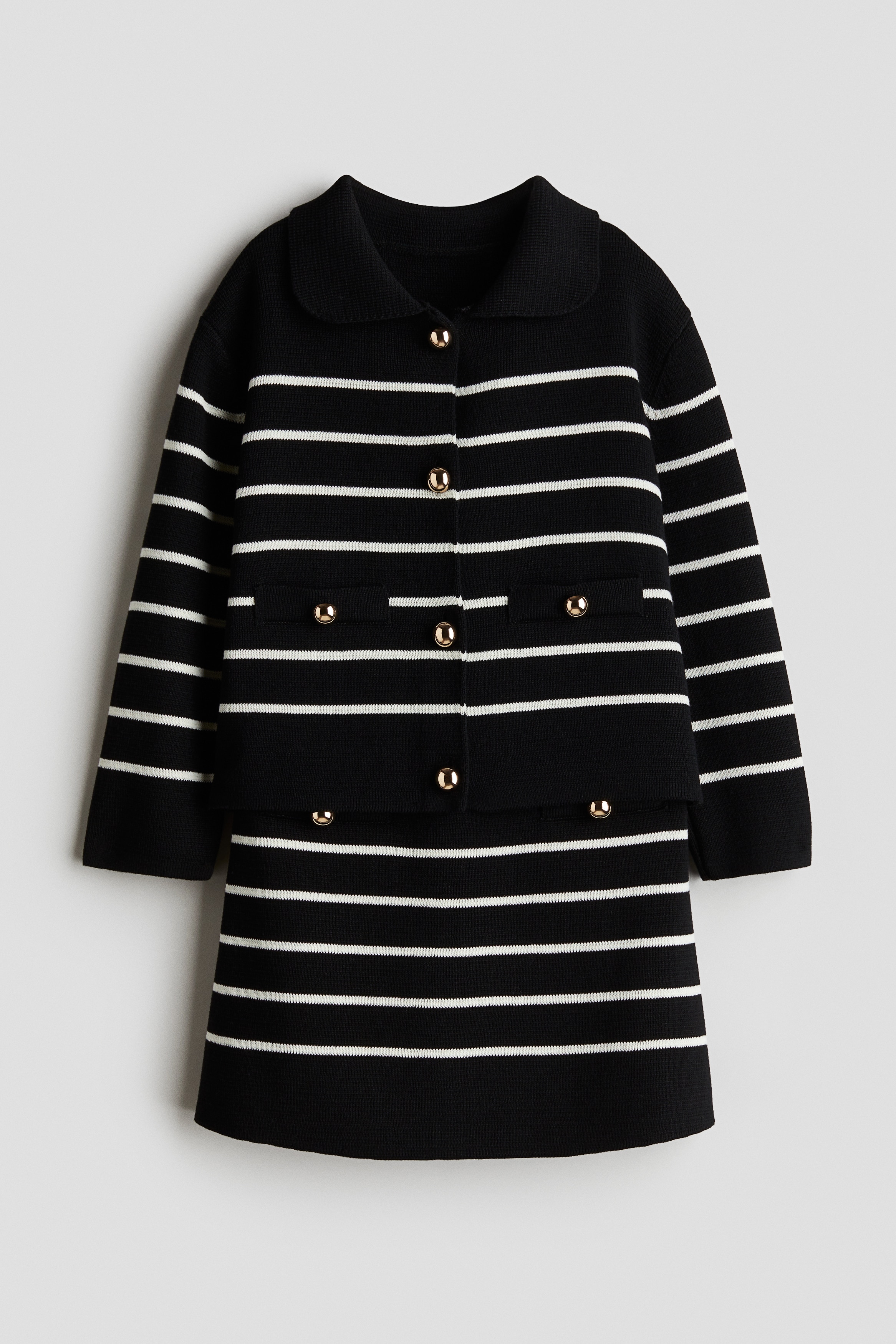 부드러운 파인니트 소재의 매치되는 가디건과 스커트 세트. 가디건 - 긴소매. 라운드 칼라. 앞면에 반짝이는 금속 버튼이 있음. 스커트 - A라인. 허릿단 히든 밴딩. 가디건과 스커트 모두 앞면의 웰트포켓 장식 부분에 금속 버튼이 있음. 이미지