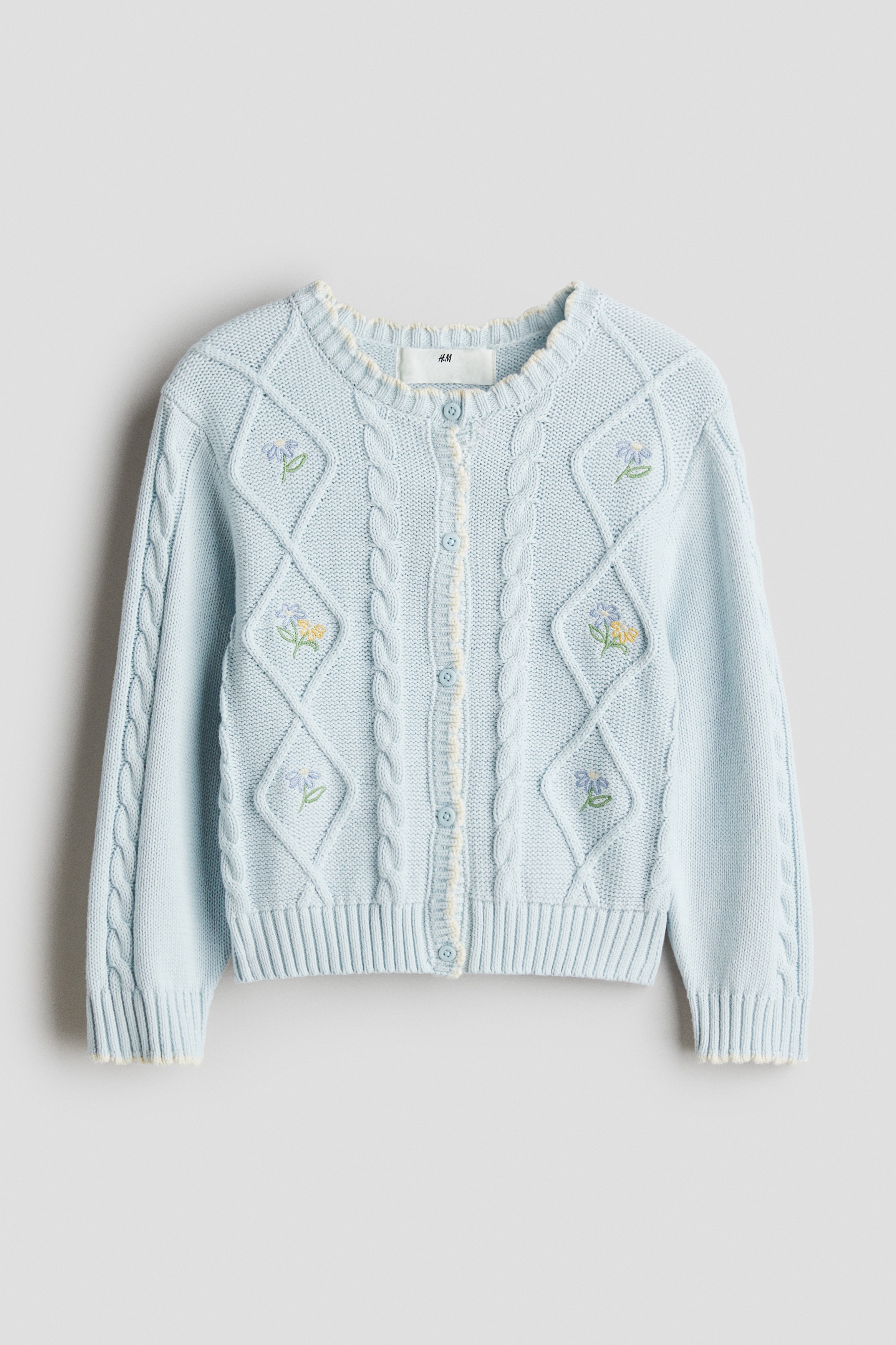 자수 장식이 있는 코튼 소재의 부드러운 케이블 니트 가디건. 스캘럽트 라인으로 마감된 옷단. 긴소매. 라운드넥. 앞면 버튼. 리브드 소맷단과 아랫단. 이미지