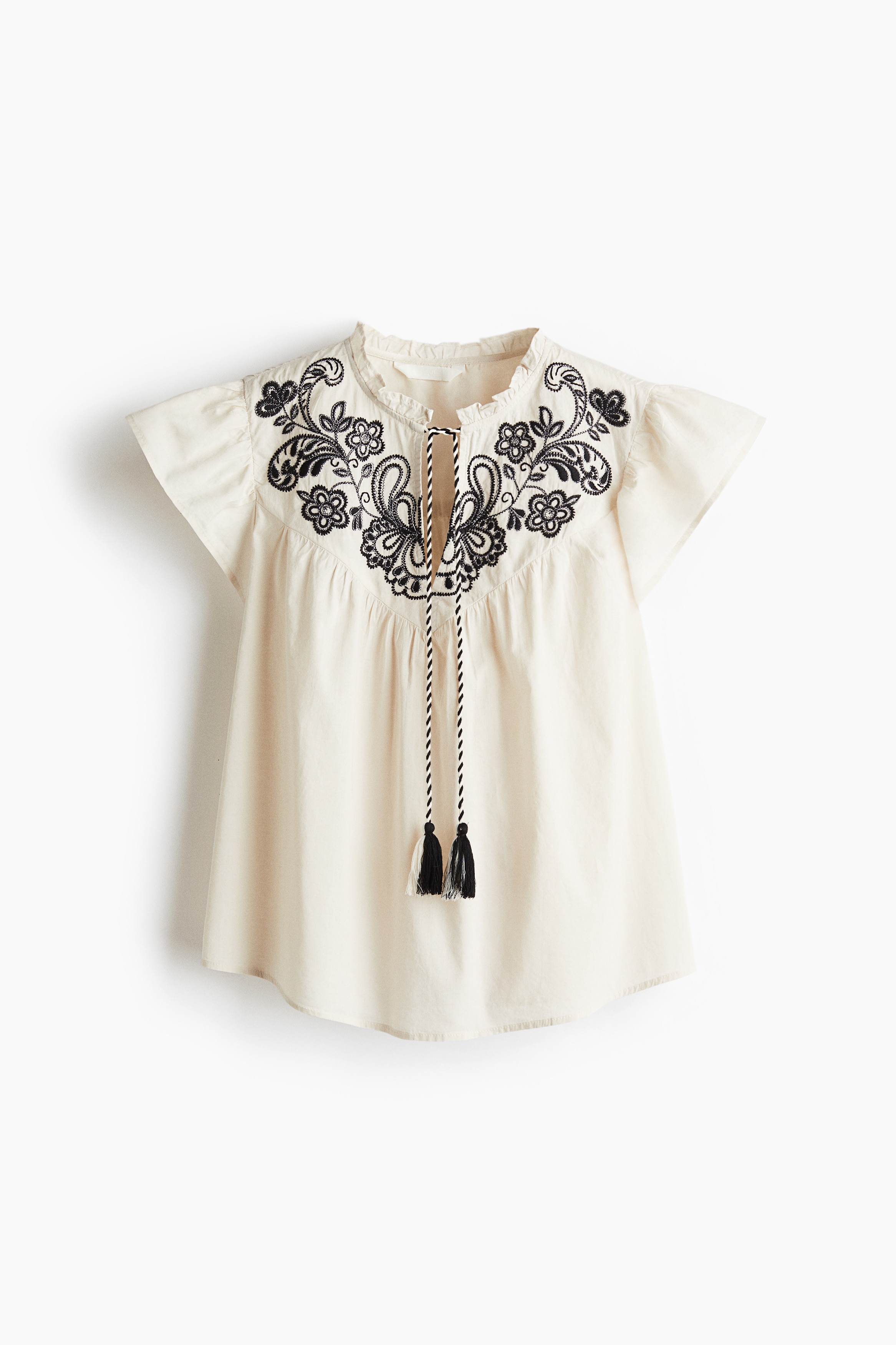 비침성 있는 코튼 소재의 블라우스. 포인트 컬러의 앞면 자수 장식. 프릴 칼라. 상단에 V자 트임과 태슬 끈이 있음. 버터플라이 소매. 주름이 잡힌 앞뒷면 요크. 세미 라운드 헴라인. 이미지
