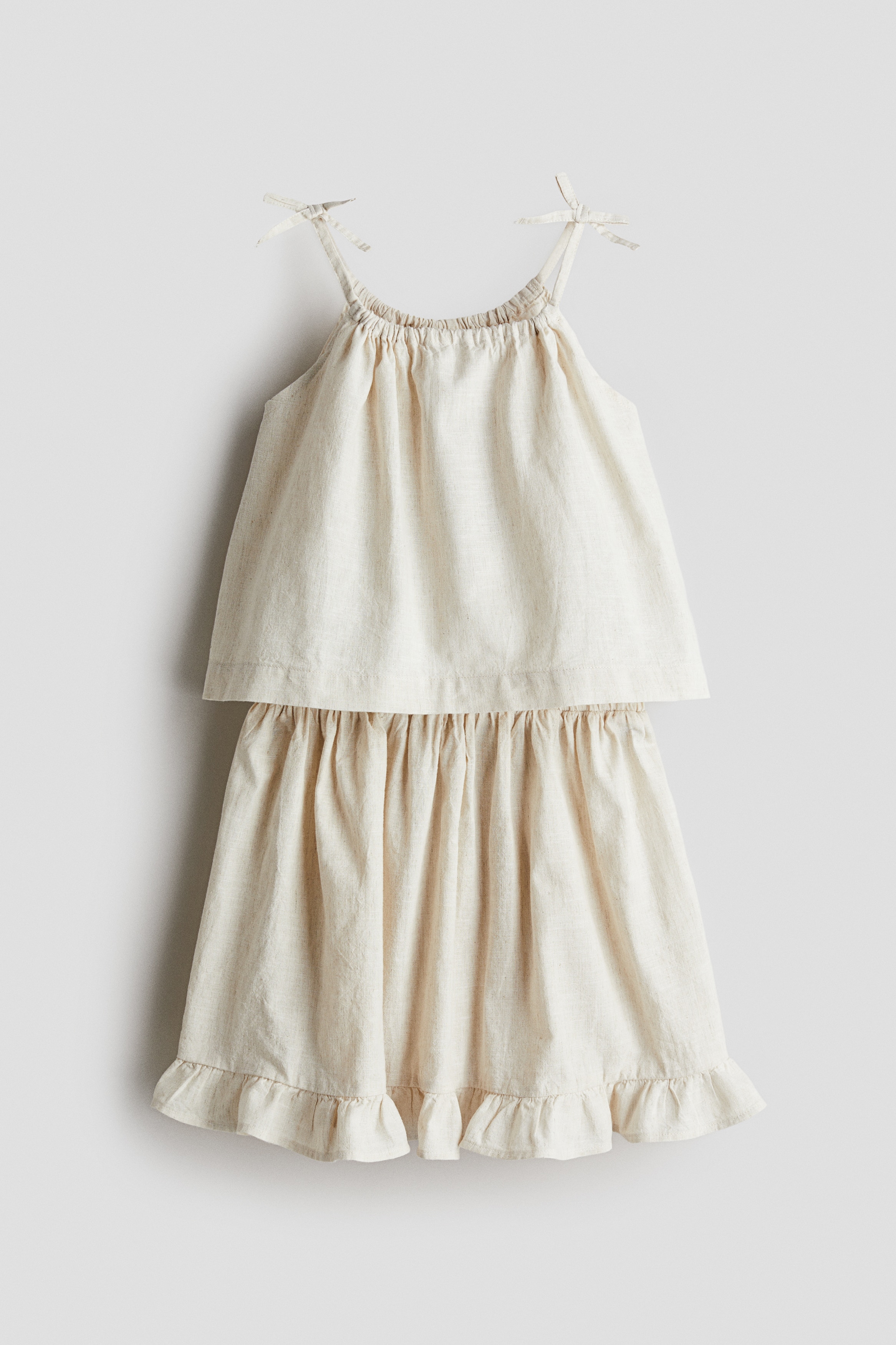 가벼운 리넨, 코튼 혼방 소재의 스트랩 탑과 스커트 세트. 탑 - 플레어 스타일. 라운드넥. 상단에 리본 장식이 부착된 가는 어깨 스트랩. 스커트 - 신축성 허릿단의 스커트. 프릴로 장식된 아랫단. 이미지