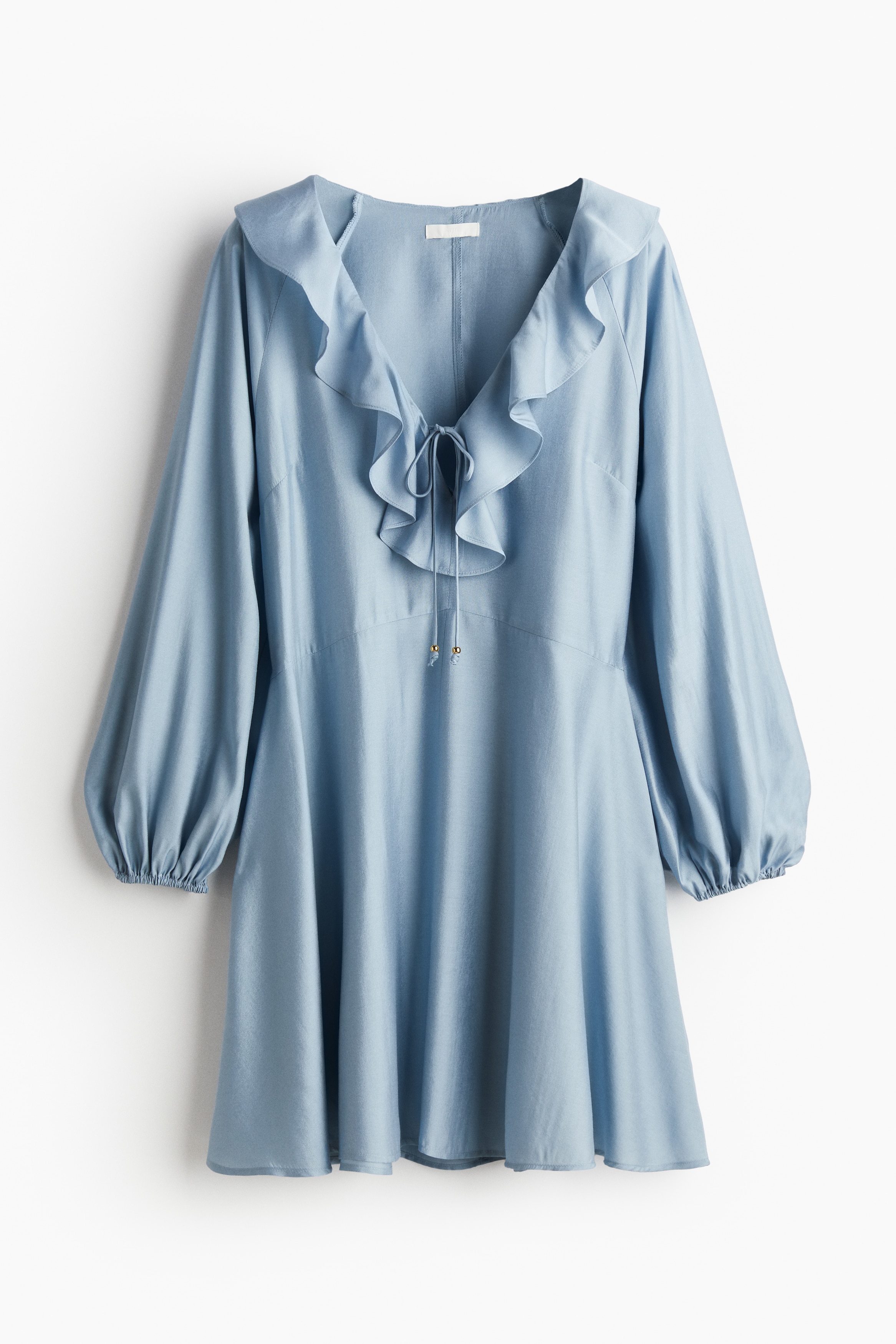 우븐 소재의 미니 원피스. 깊게 파인 V넥이 플라운스로 장식되었으며, 앞면에 가는 끈이 있음. 래글런 스타일의 긴 벌룬 소매. 신축성 소맷단. 플레어 라인의 하단. 안감 생략. 이미지