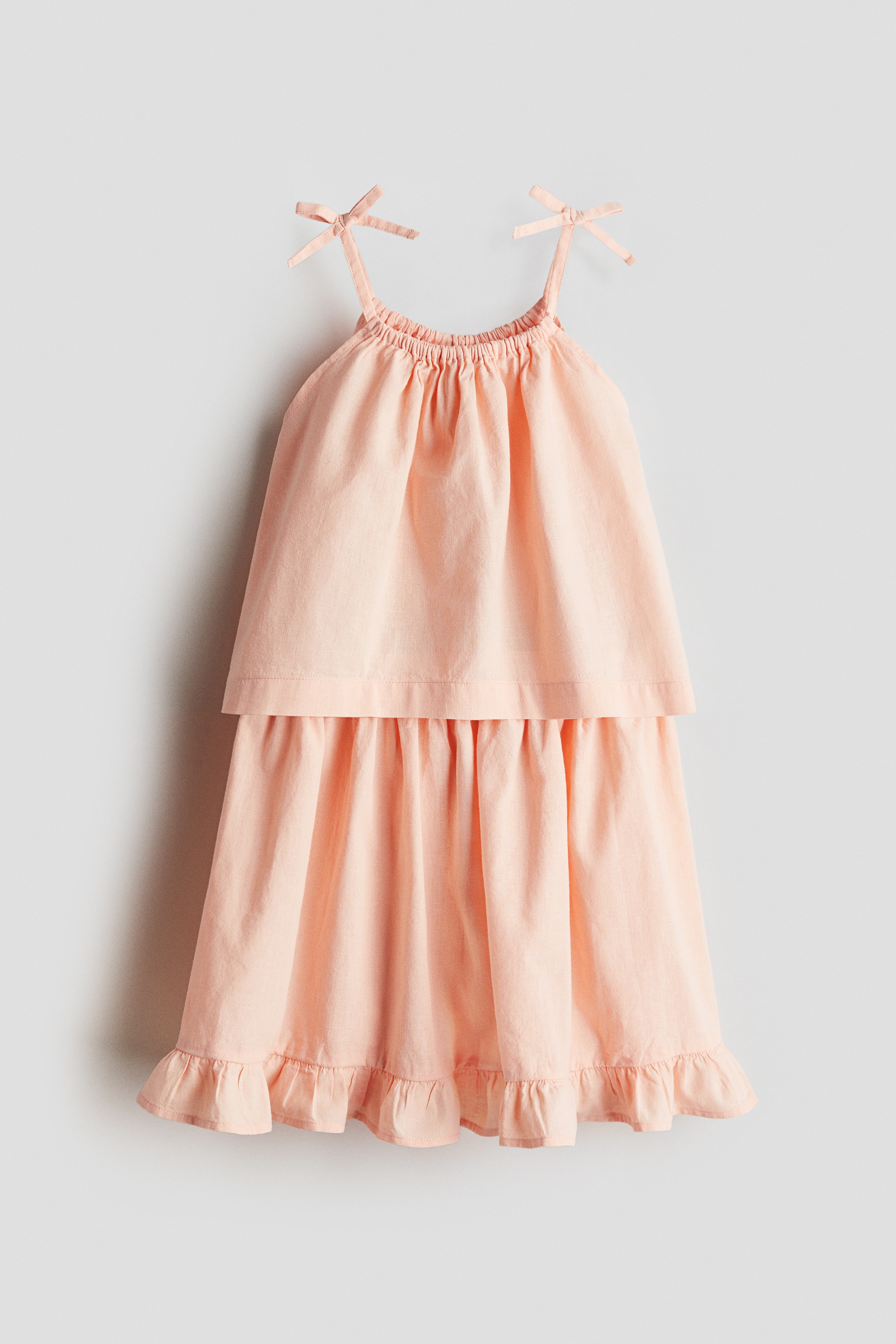 가벼운 리넨, 코튼 혼방 소재의 스트랩 탑과 스커트 세트. 탑 - 플레어 스타일. 라운드넥. 상단에 리본 장식이 부착된 가는 어깨 스트랩. 스커트 - 신축성 허릿단의 스커트. 프릴로 장식된 아랫단. 이미지