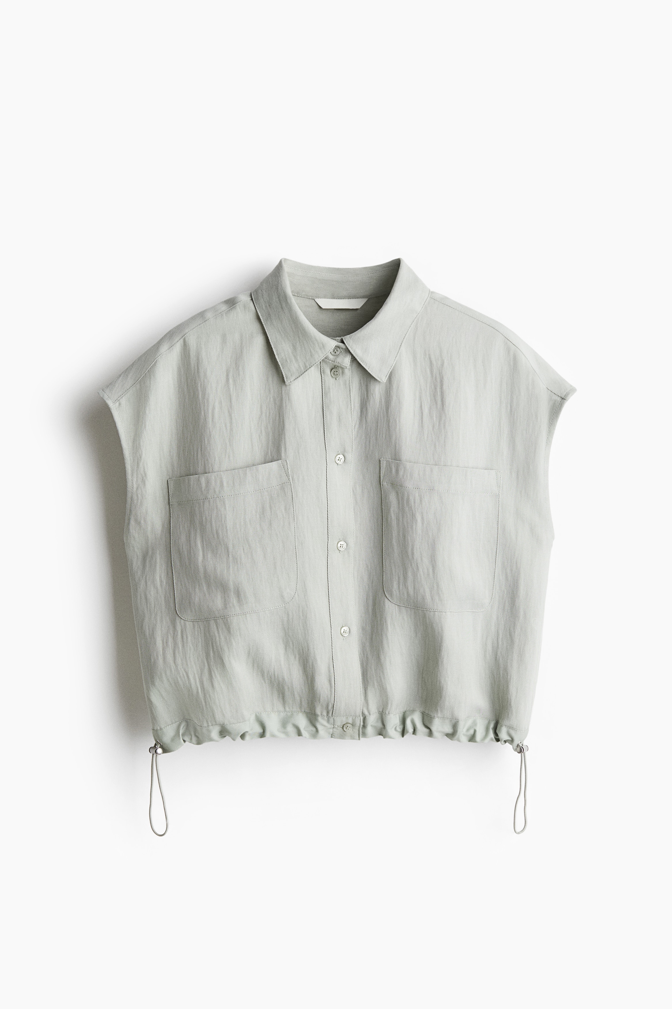 리오셀이 혼방된 트윌 소재의 슬리브리스 셔츠. 칼라와 앞면 버튼이 있음. 가슴 포켓. 플리츠가 잡힌 뒷면 요크 디테일. 양쪽 옆면에 신축성 드로스트링과 코드락이 있는 아랫단. 이미지