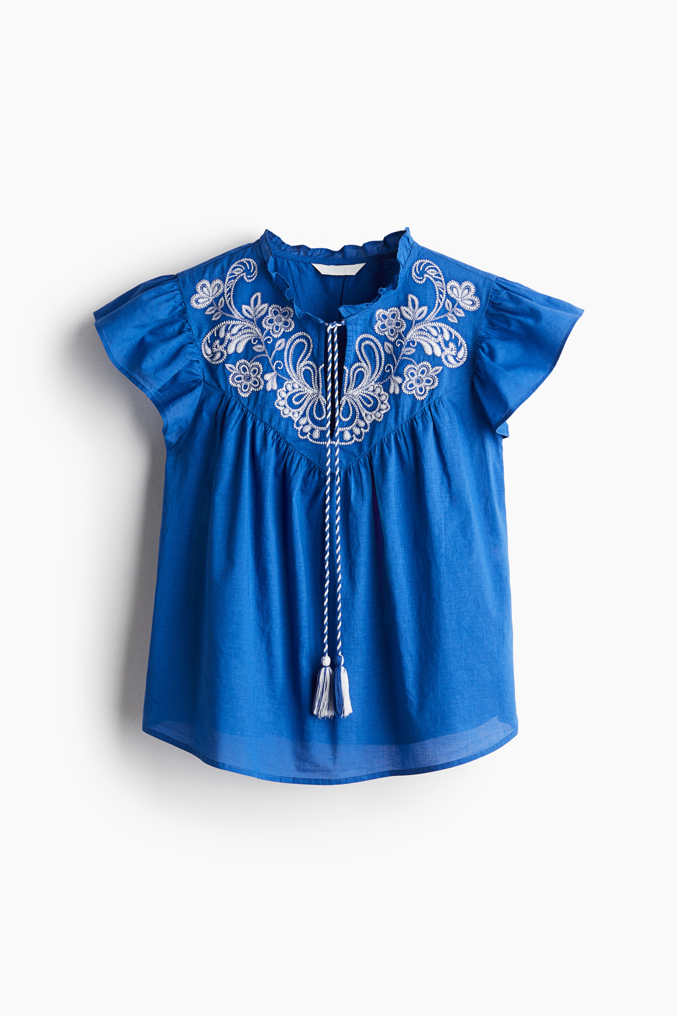 비침성 있는 코튼 소재의 블라우스. 포인트 컬러의 앞면 자수 장식. 프릴 칼라. 상단에 V자 트임과 태슬 끈이 있음. 버터플라이 소매. 주름이 잡힌 앞뒷면 요크. 세미 라운드 헴라인. 이미지