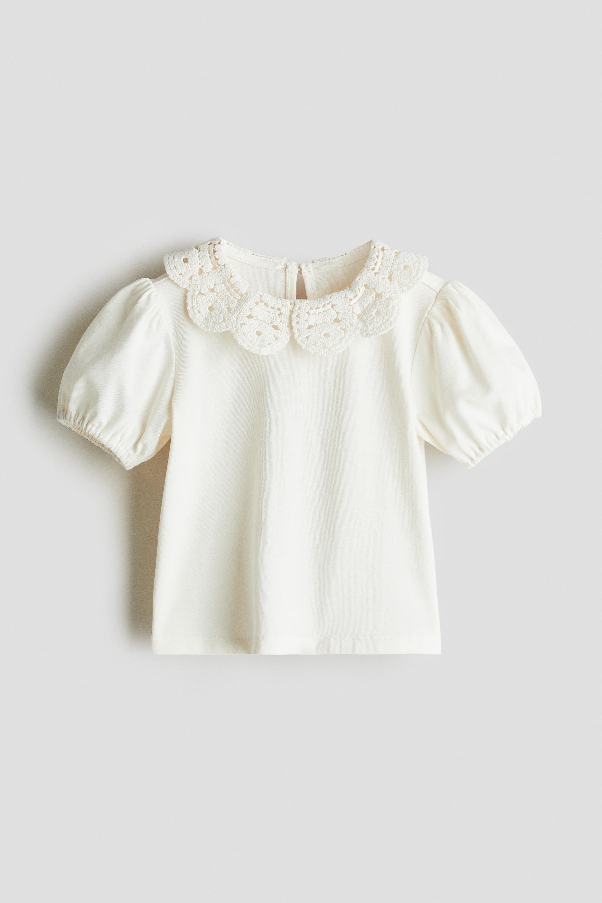 부드러운 코튼 저지 소재의 탑. 크로셰 느낌의 니트를 사용한 스캘럽트 라인의 라운드 칼라. 라운드넥. 뒷면에 키홀 트임과 버튼이 있음. 퍼프 소매. 신축성 소맷단. 이미지