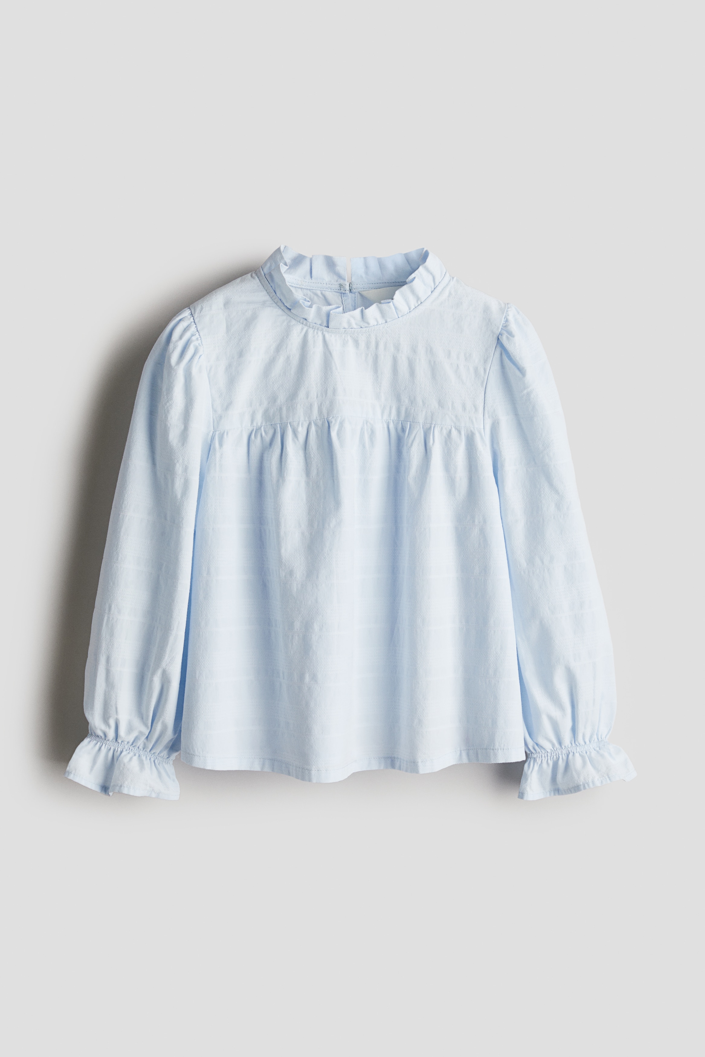 코튼 소재의 부드러운 자카드 블라우스. 프릴로 마감된 라운드넥. 목 뒷면에 키홀 트임과 버튼이 있음. 주름이 잡힌 앞뒷면 요크. 손목 부분이 가늘게 밴딩처리된 퍼프 스타일의 긴소매. 프릴 소맷단. 이미지