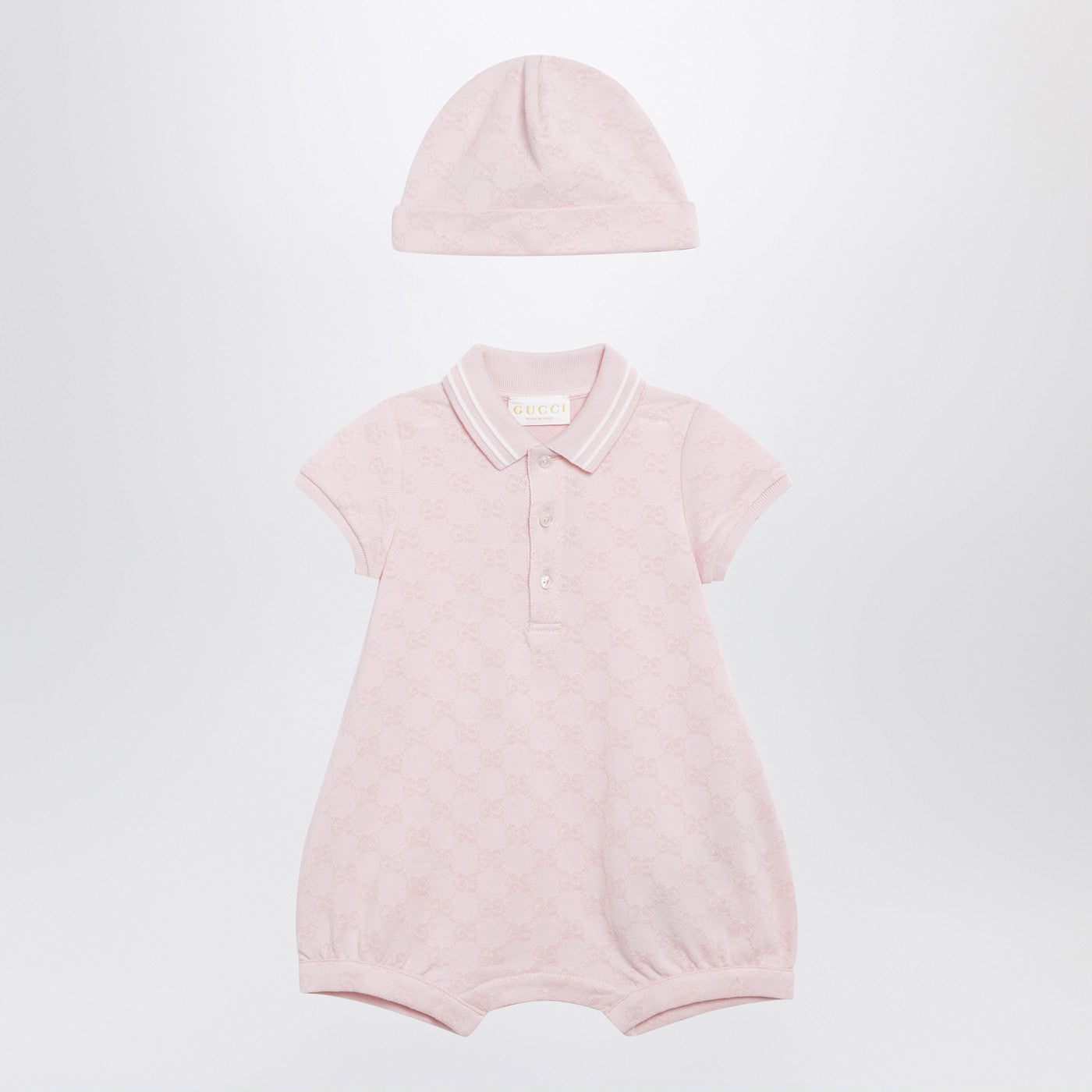 구찌의 라이트 핑크 GG 피케 코튼 투피스 세트는 스트라이프 폴로 스타일 칼라, 짧은 소매, 신축성 있는 밑단, 골지 트리밍, 숨겨진 스냅 버튼 클로저가 특징인 롬퍼와 매칭되는 모자로 구성되어 있습니다. 이미지